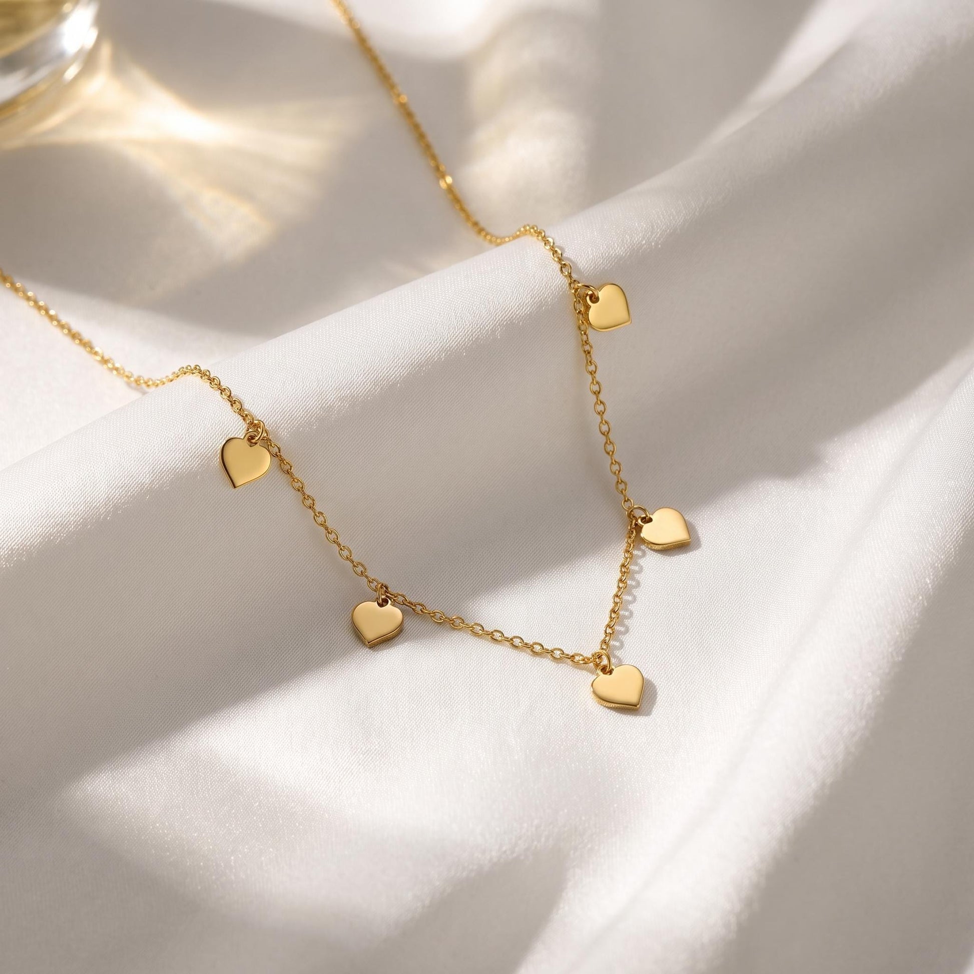 A delicate gold necklace with small heart-shaped charms dangles from a white fabric surface, creating a romantic and elegant appearance.