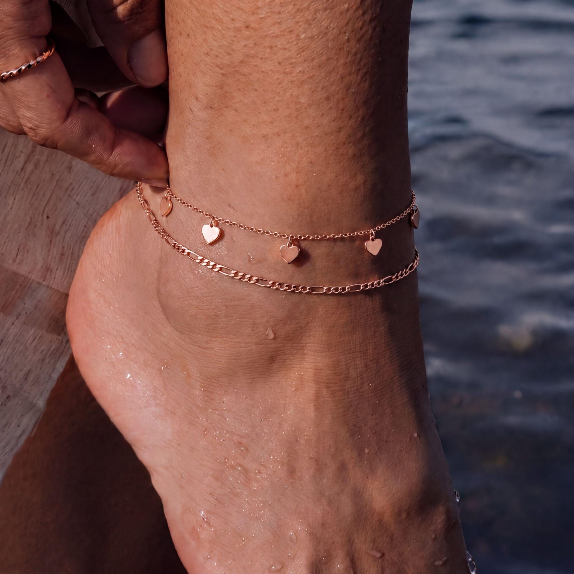 A close-up view of a person's ankle wearing a delicate gold-colored ankle bracelet with small heart-shaped charms.