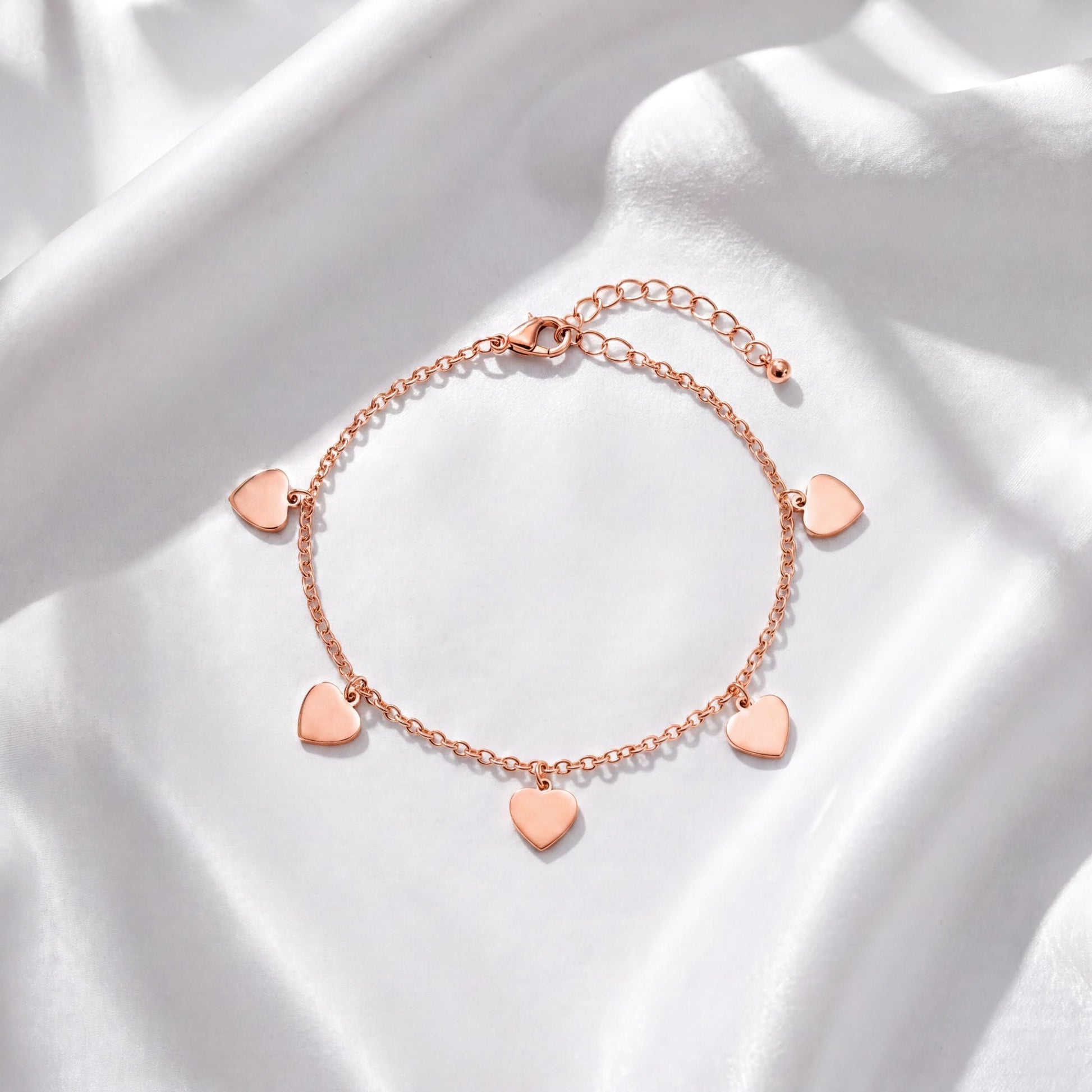 A delicate gold-colored bracelet with small heart-shaped charms attached to it, resting on a white fabric background.