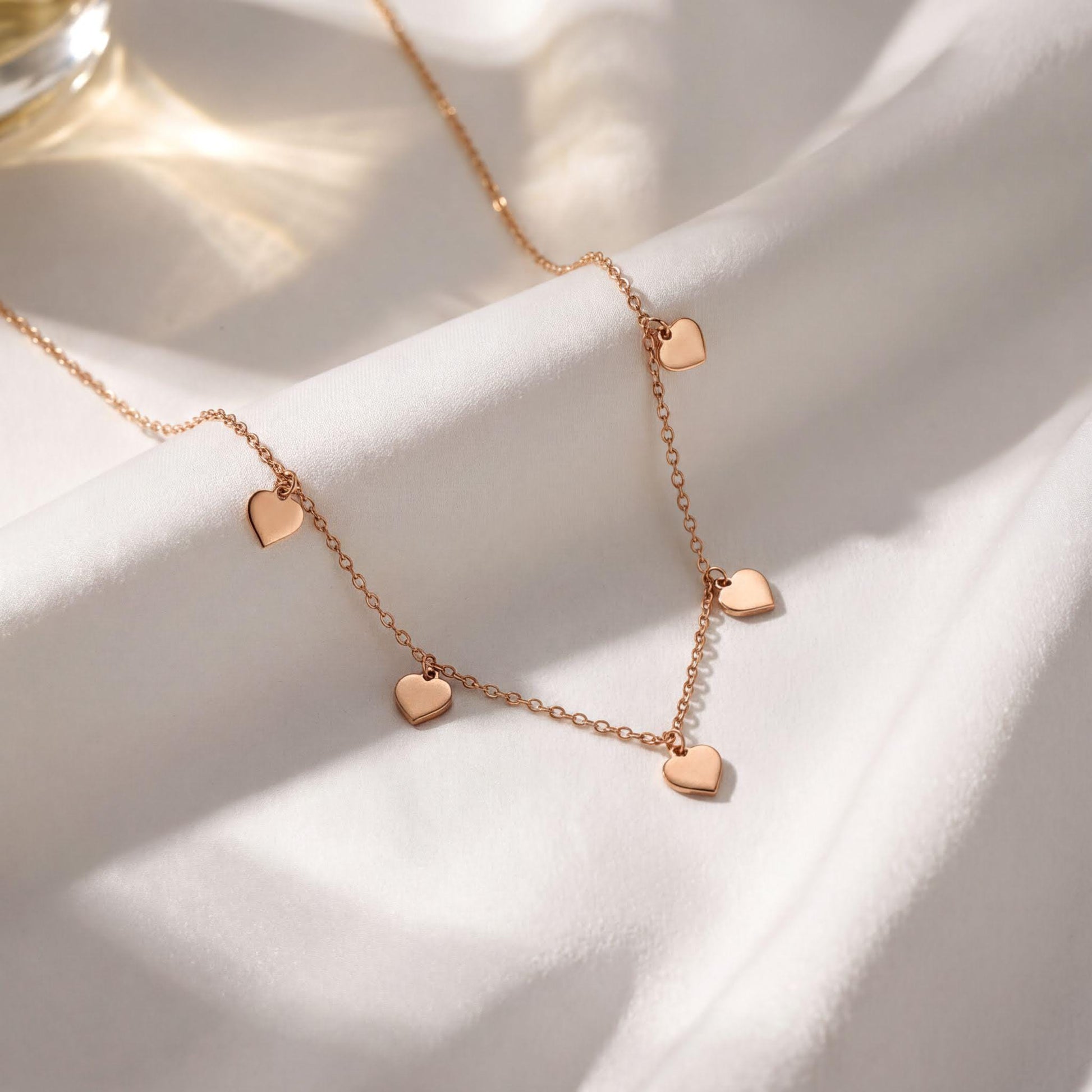 A delicate gold necklace with small heart-shaped charms dangles from a white fabric surface, creating a romantic and elegant display.