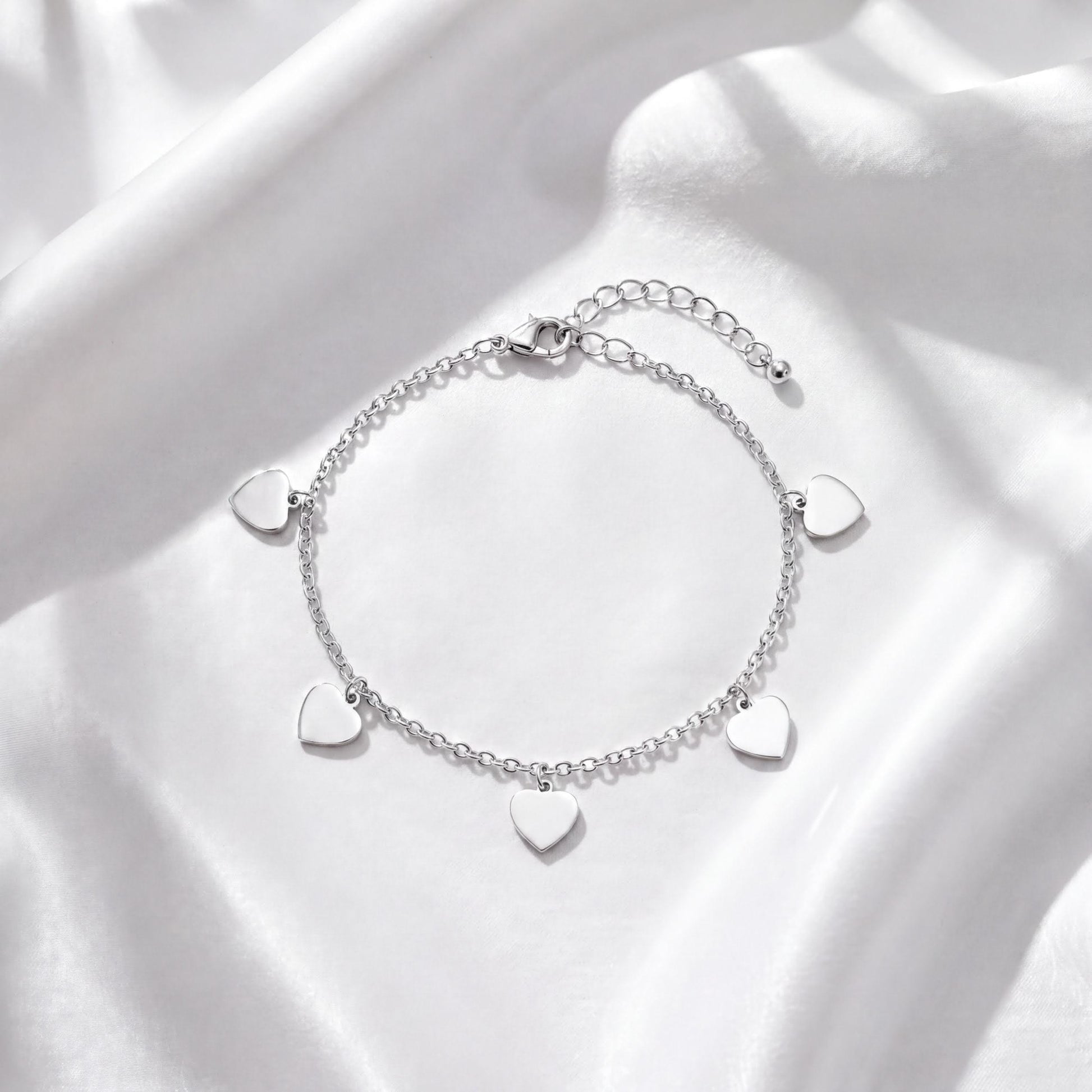 A silver bracelet with heart-shaped charms is displayed on a white fabric background.