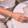 A person's hand wearing a gold bracelet is resting on a table with a glass of water and a lemon slice.