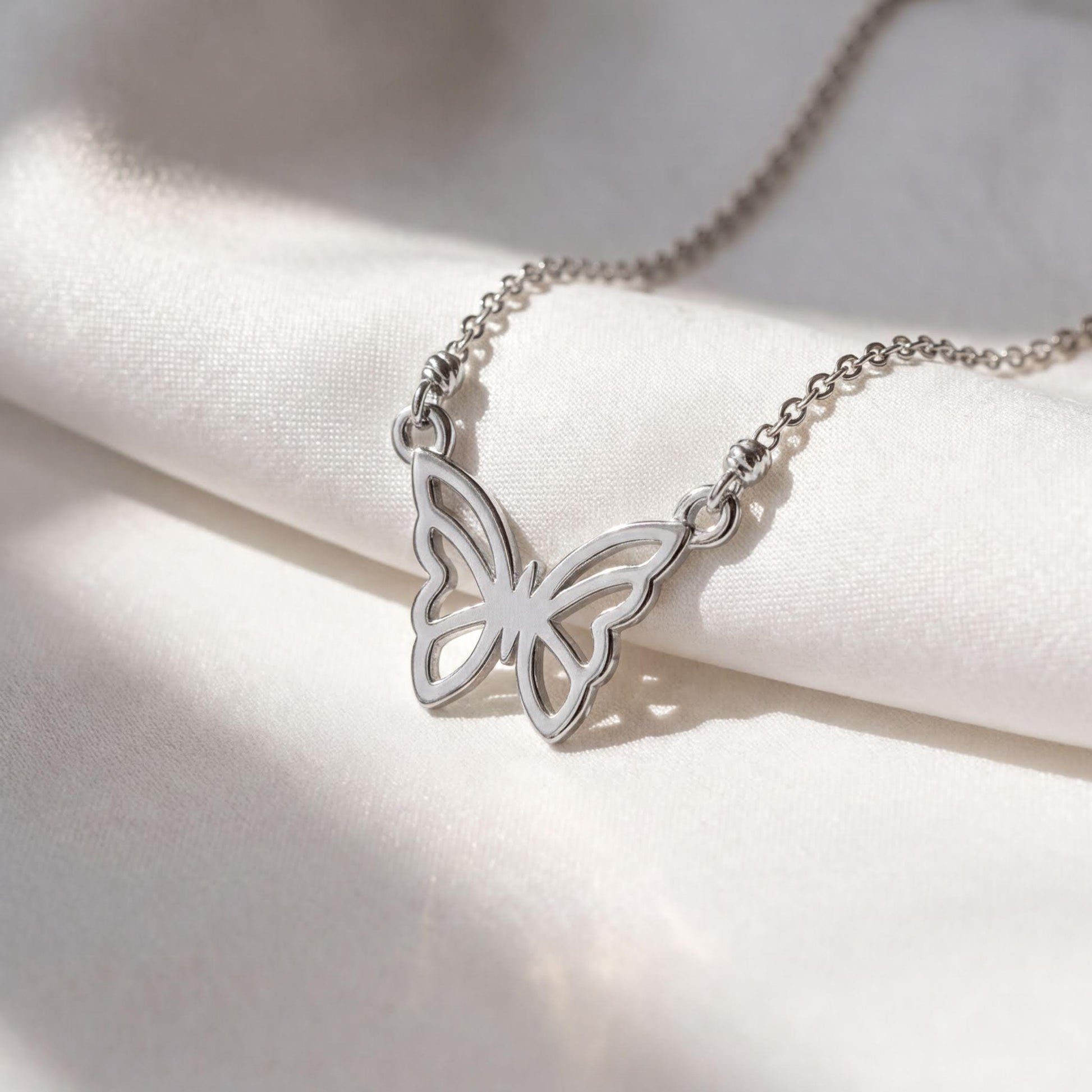 A silver necklace with a butterfly pendant is displayed on a white fabric surface.