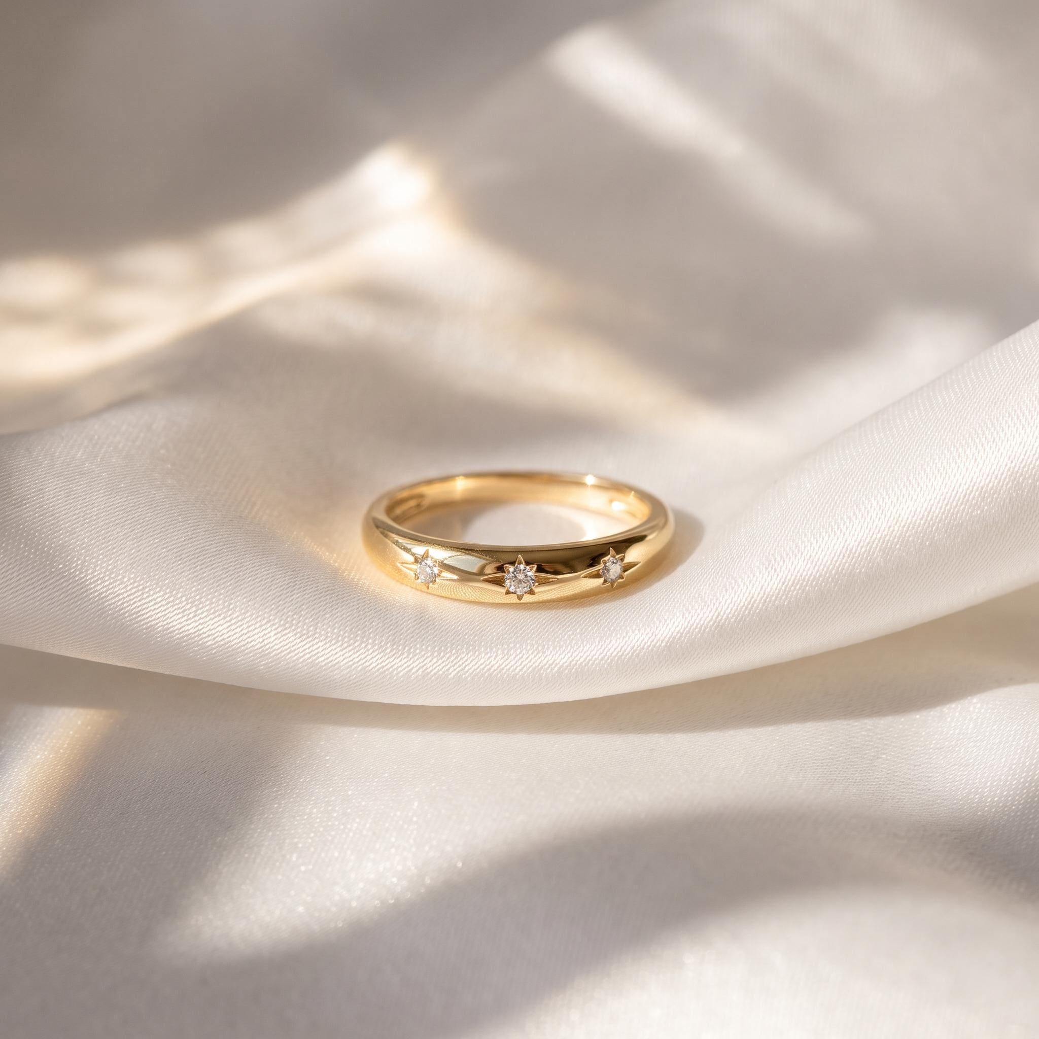 A gold ring with three small diamonds is displayed on a white fabric surface, with sunlight casting shadows and highlighting the details of the ring.