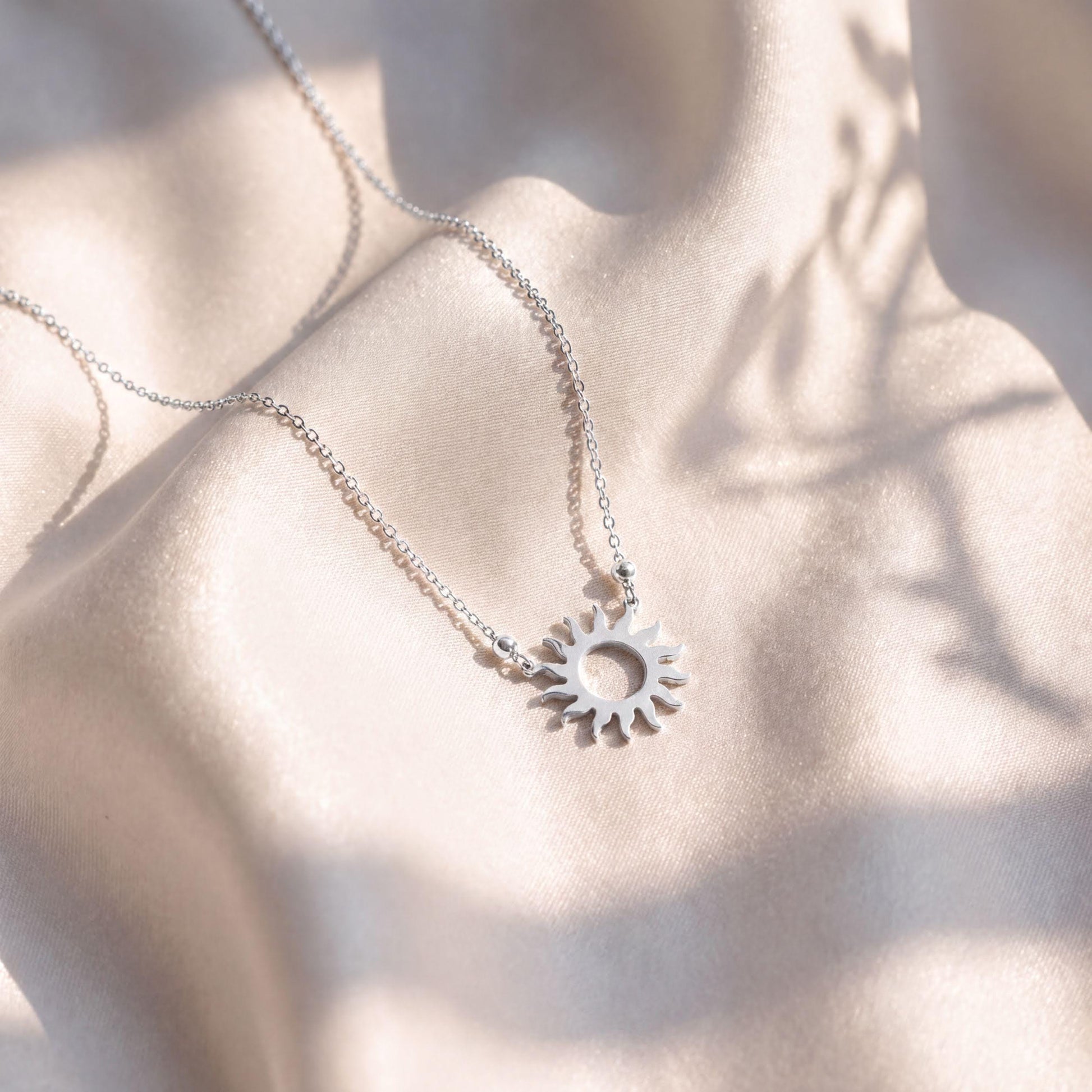 A delicate silver necklace with a pendant featuring a sun-like design rests on a soft, light-colored fabric.