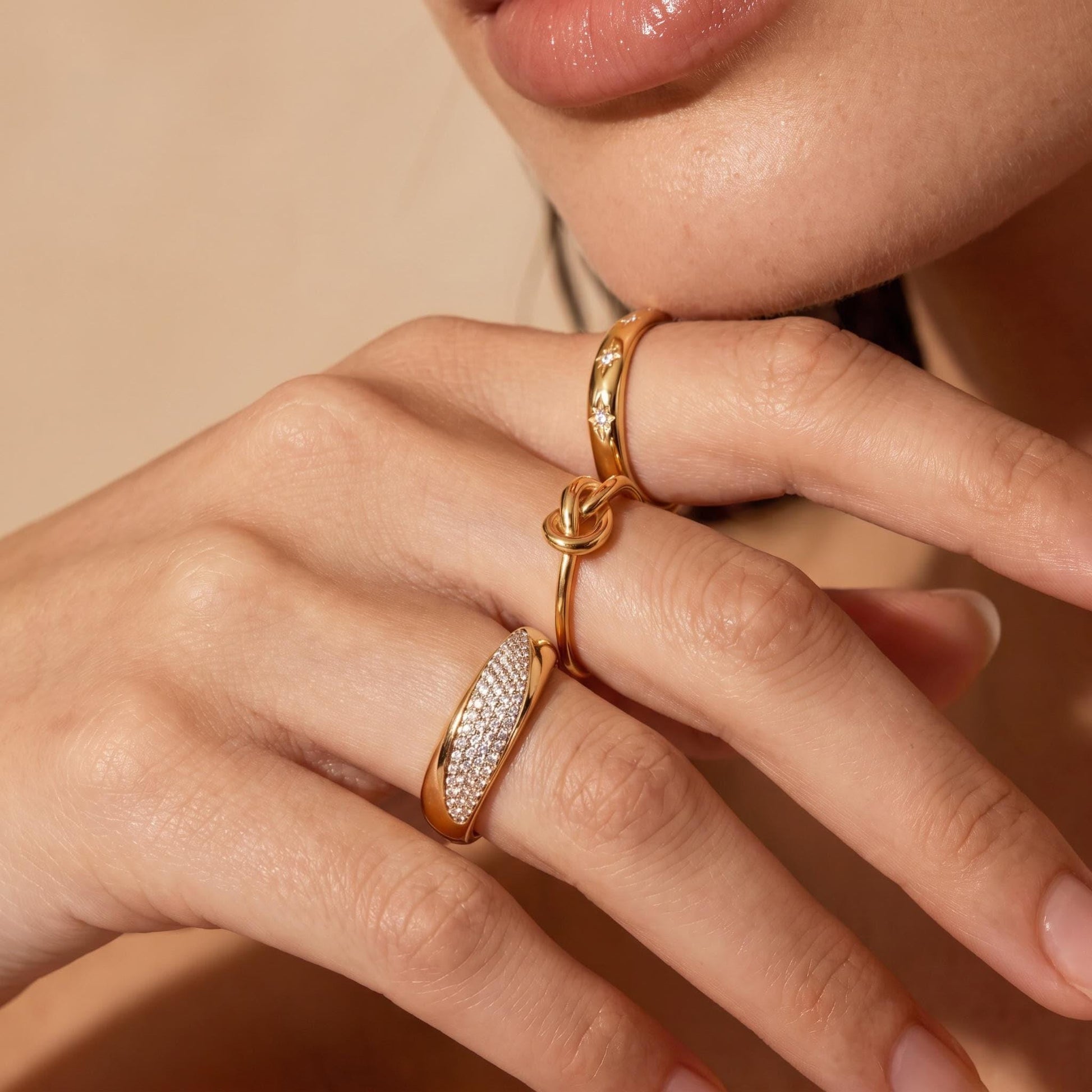 A close-up of a person's hand wearing multiple gold rings, including a ring with a diamond-encrusted band and another ring with a knotted band.