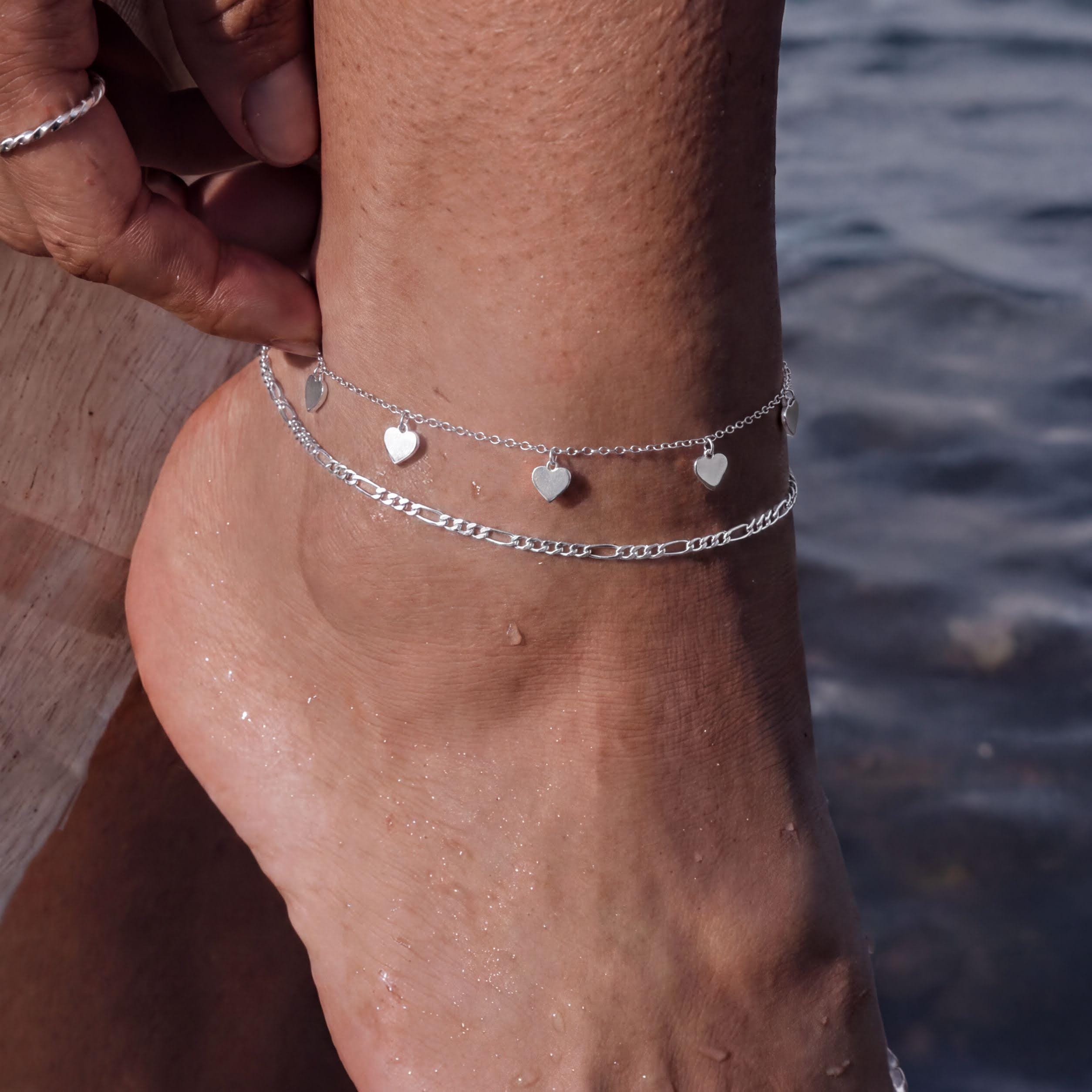 A close-up view of a person's ankle wearing a silver ankle bracelet with heart-shaped charms.