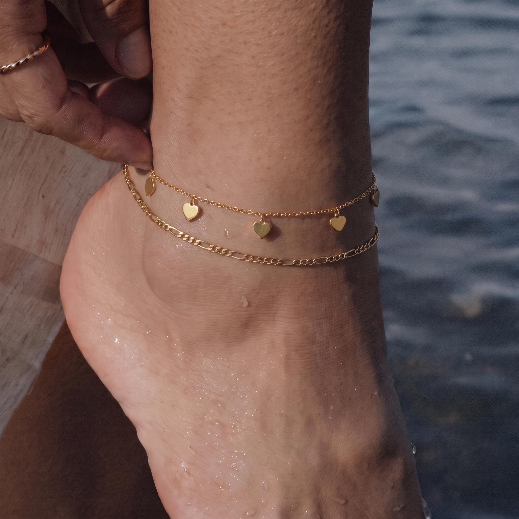 A close-up of a person's ankle wearing a gold-colored ankle bracelet with small heart-shaped charms.