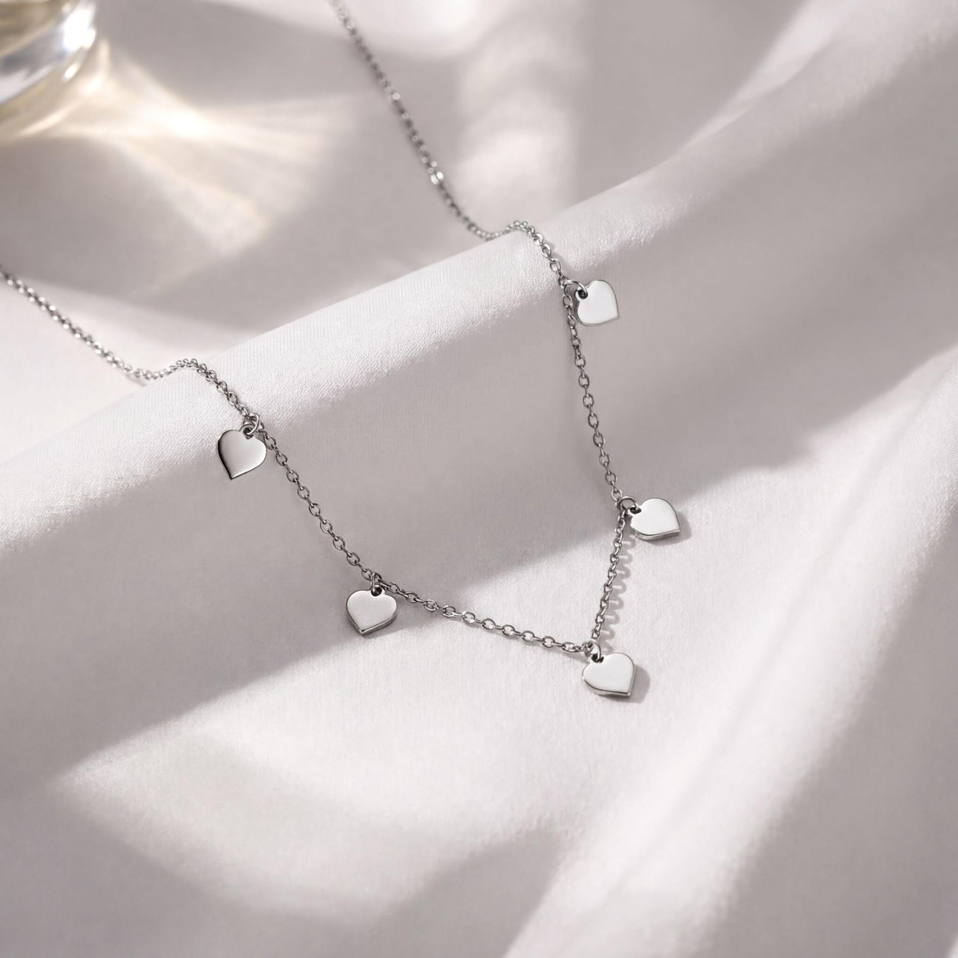 A delicate silver necklace with small heart-shaped charms dangles from a chain, resting on a white fabric surface.