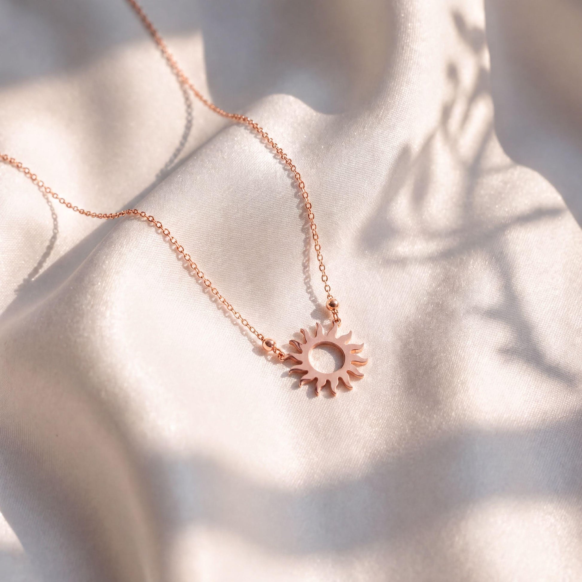 A delicate necklace with a sun-shaped pendant rests on a white fabric surface, casting a shadow on the fabric.