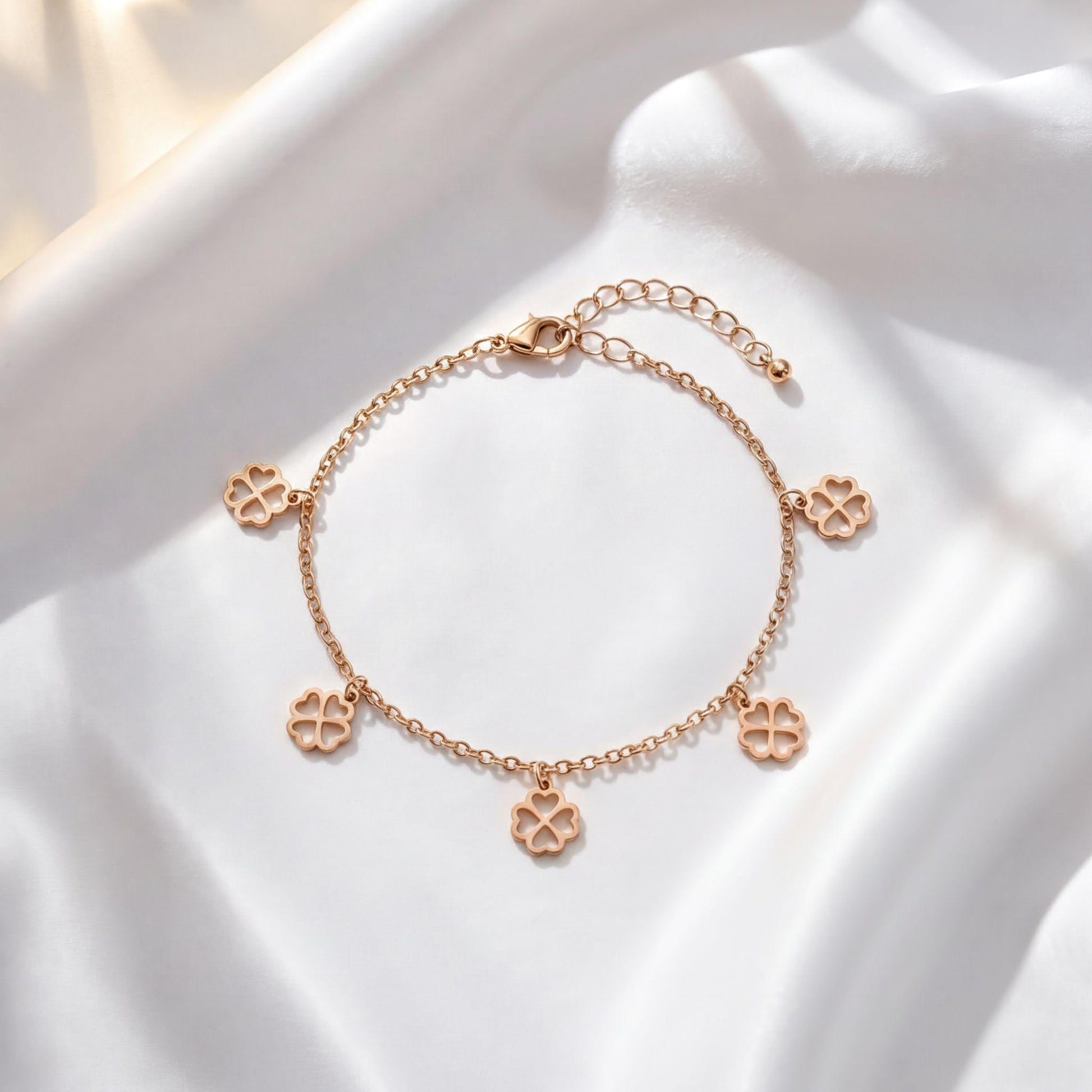 A delicate gold-colored bracelet with four charms, each featuring a four-leaf clover design, is displayed on a white fabric surface.