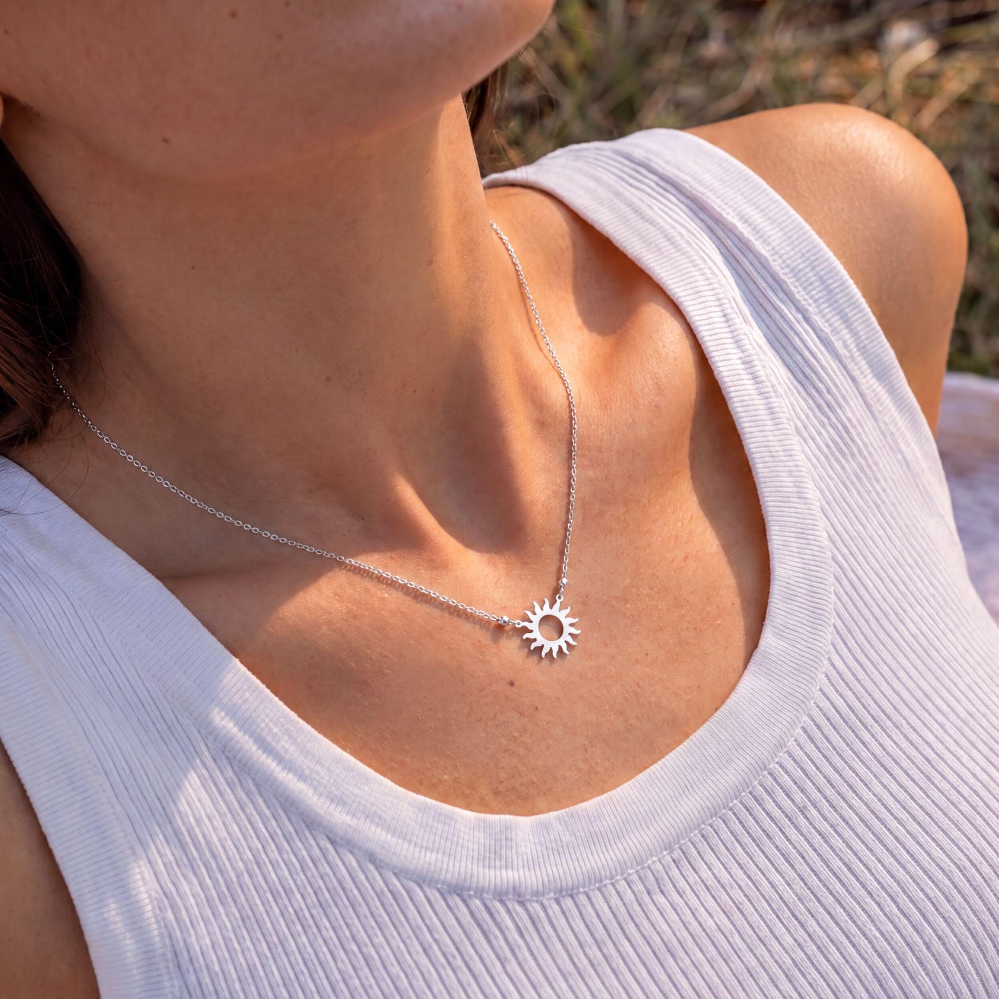 A woman wearing a white tank top and a silver necklace with a sun pendant is shown in the image.