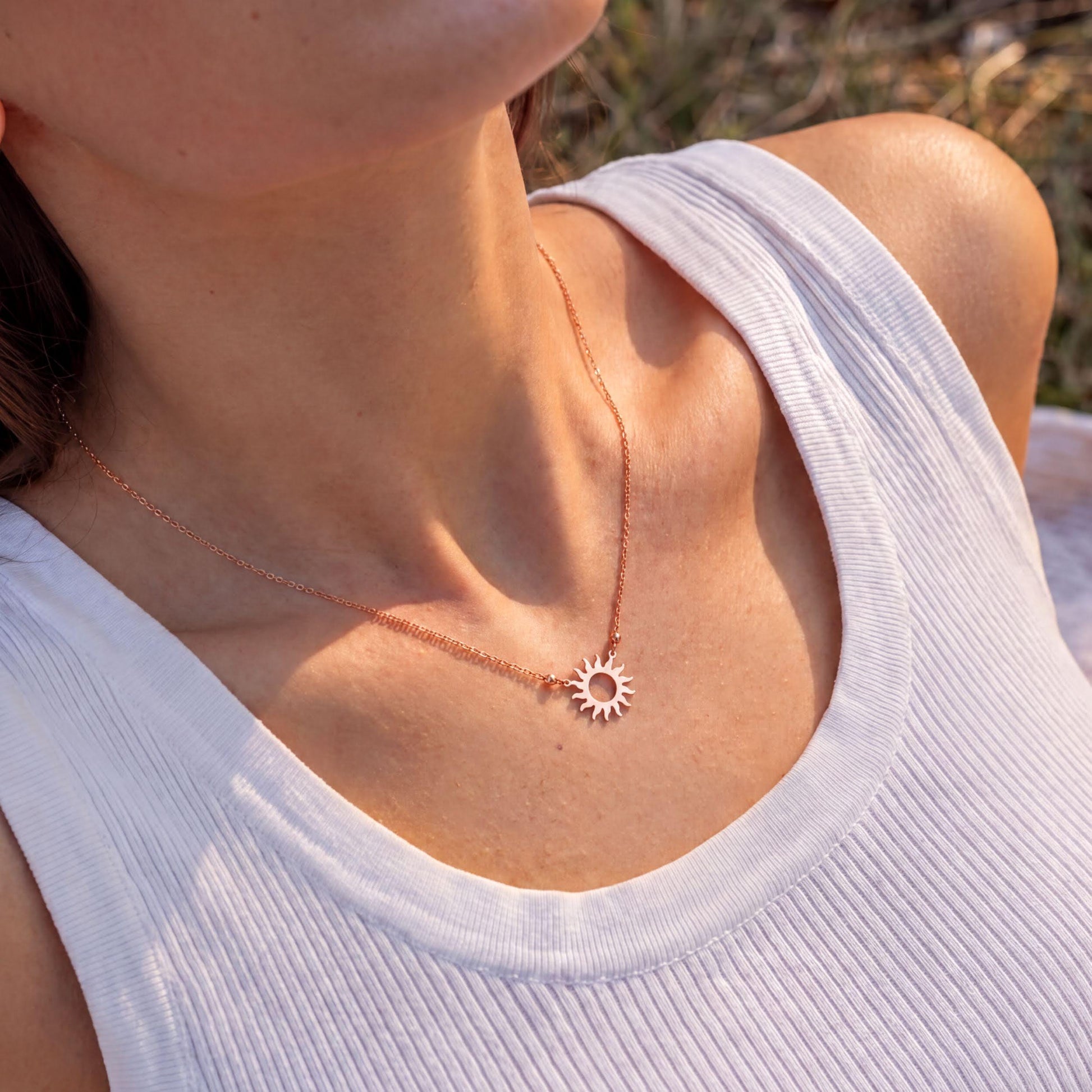 A woman wearing a white tank top and a necklace with a sun pendant is shown in the image.