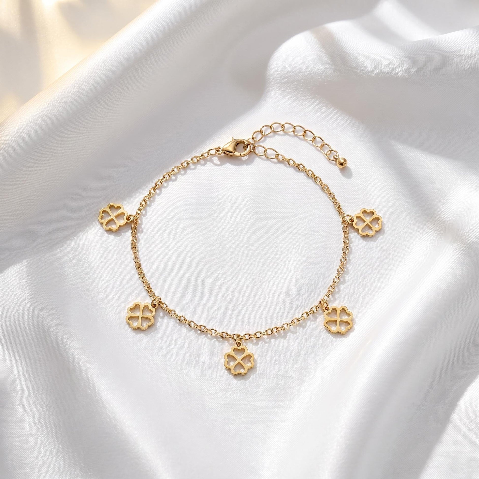 A delicate gold-colored bracelet with four charms, each featuring a four-leaf clover design, is displayed on a white fabric surface.