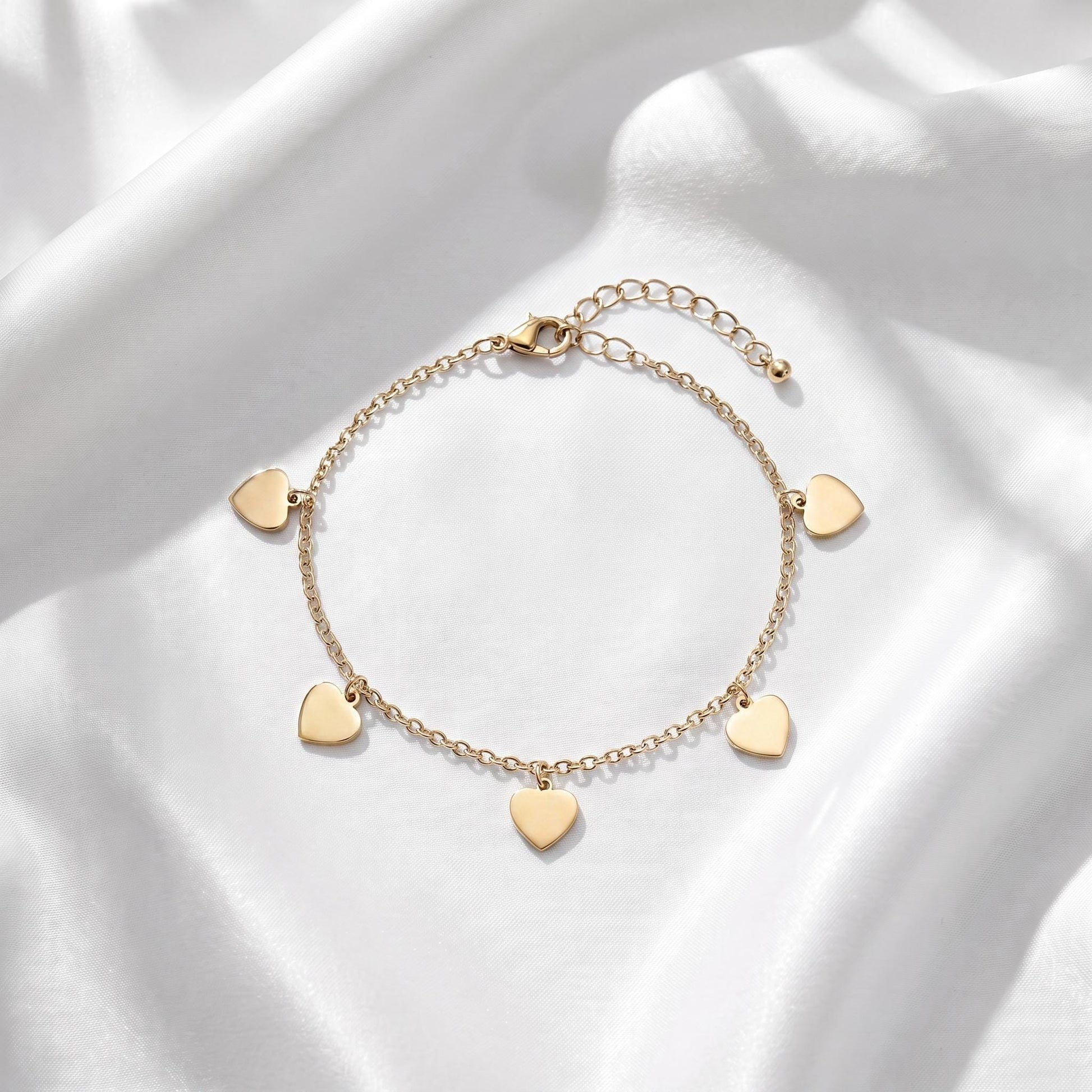 A delicate gold-colored bracelet with small heart-shaped charms attached to it, resting on a white fabric background.