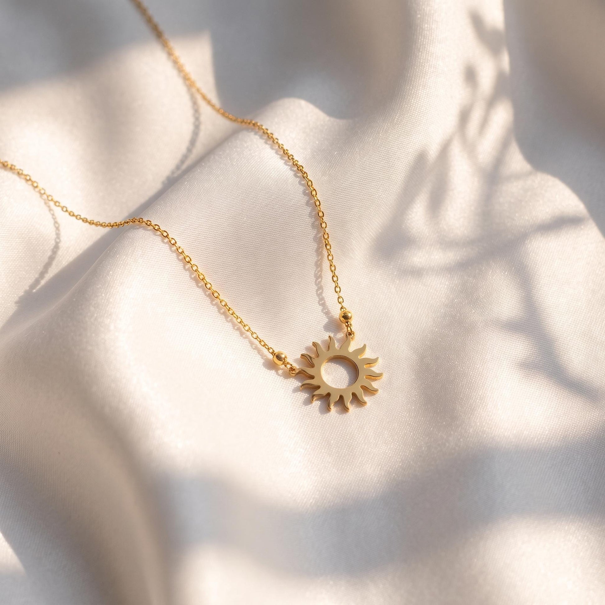 A delicate gold necklace with a sun-shaped pendant rests on a white fabric surface, casting a shadow on the fabric.
