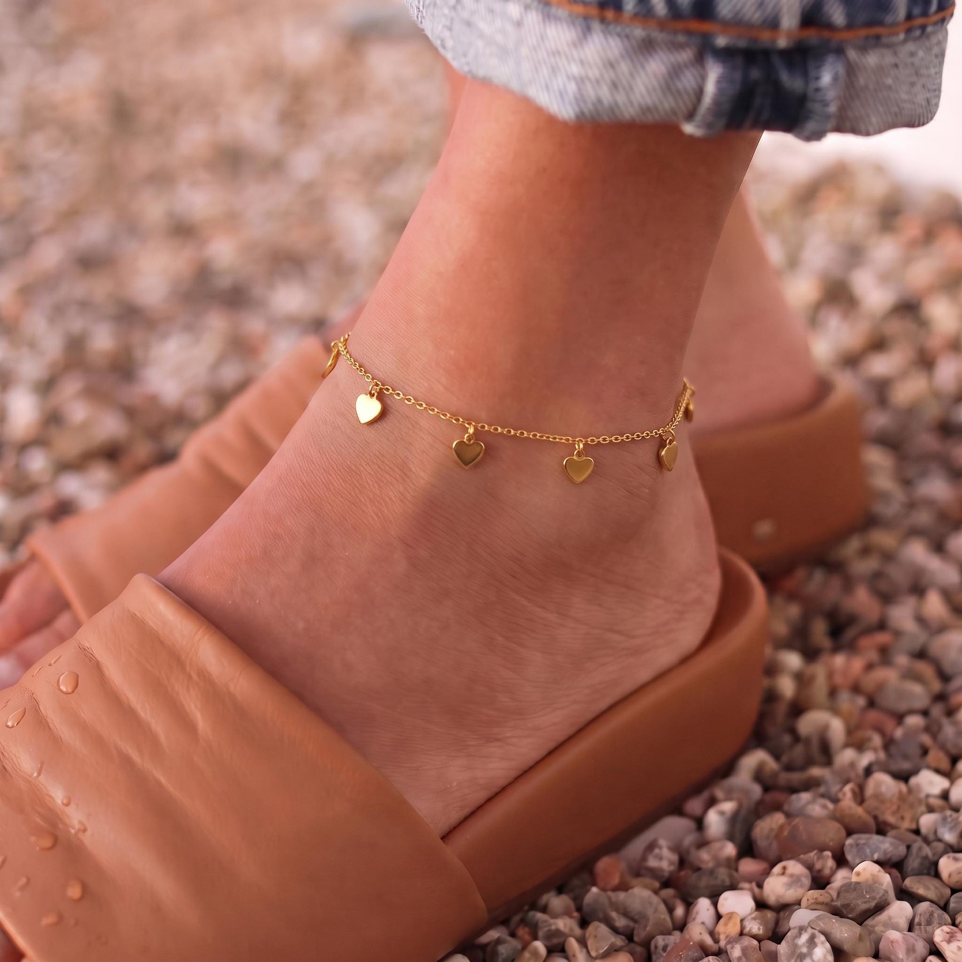 A close-up of a person's ankle wearing a gold ankle bracelet with heart-shaped charms.