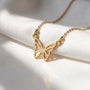 A delicate gold-colored necklace with a butterfly pendant is displayed on a white fabric surface, with sunlight casting a shadow on the fabric.