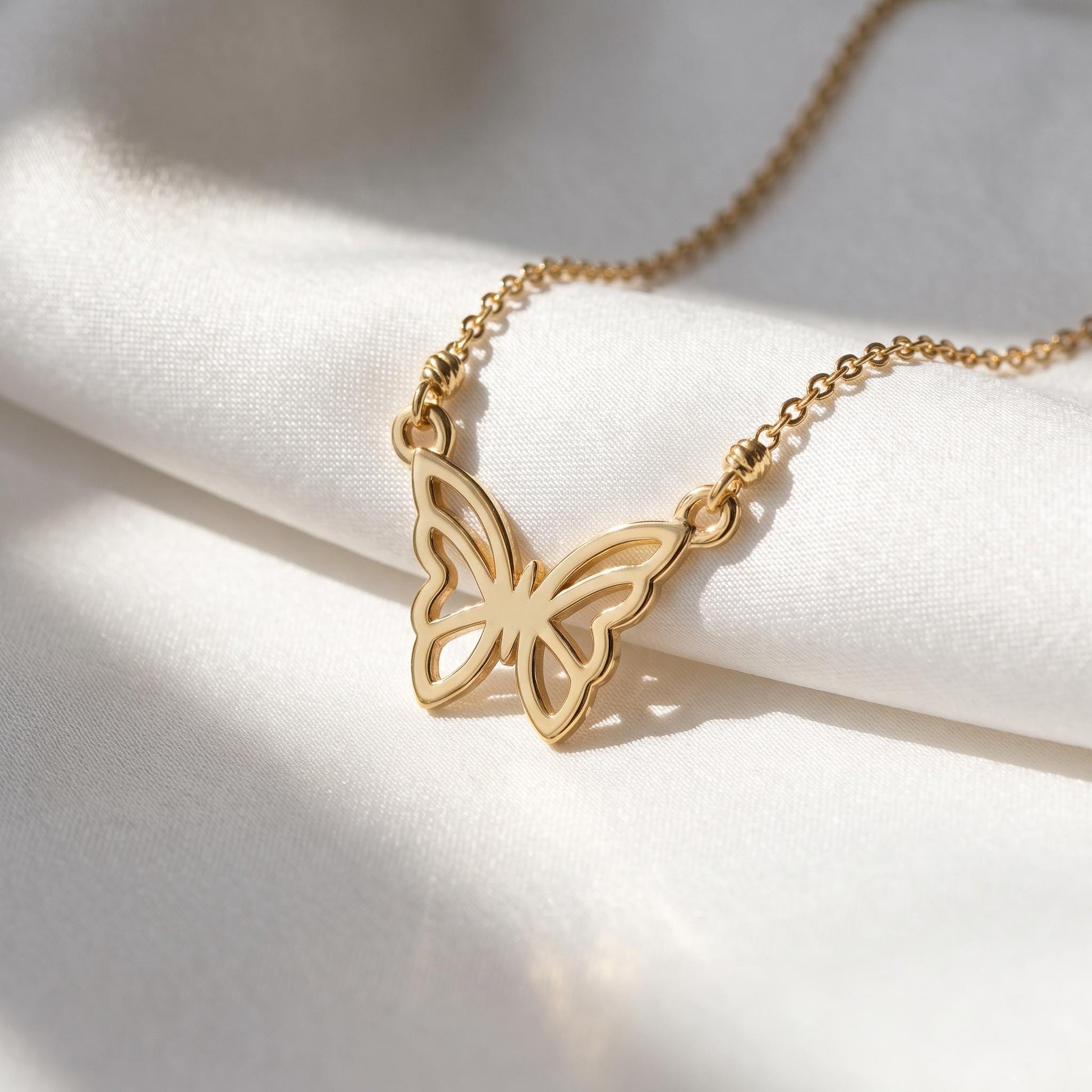 A delicate gold-colored necklace with a butterfly pendant is displayed on a white fabric surface, with sunlight casting a shadow on the fabric.